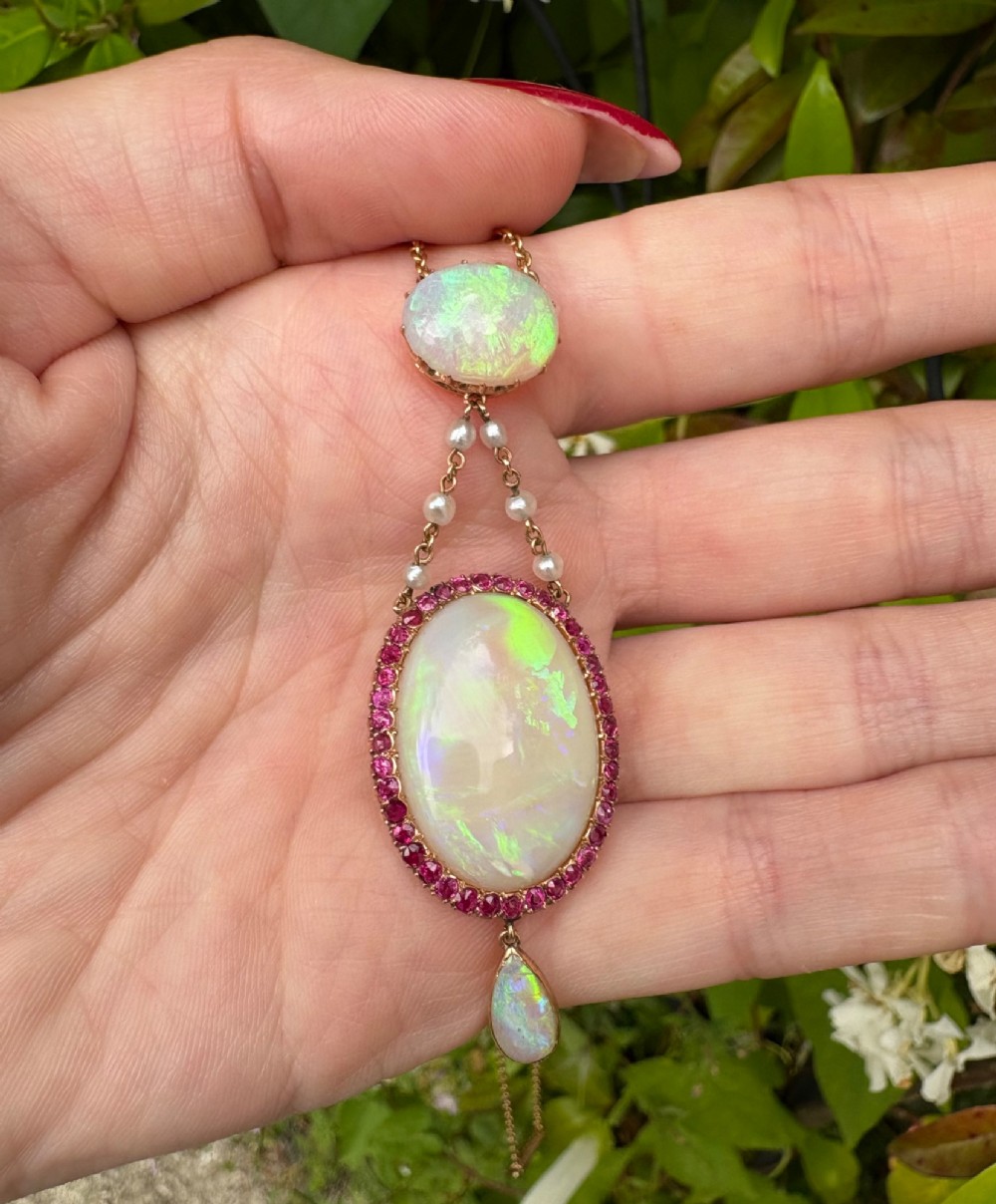 An antique Victorian 15ct yellow gold lavalier pendant set with two oval cabochon opals, a halo of rubies, and seed pearl connectors. View V