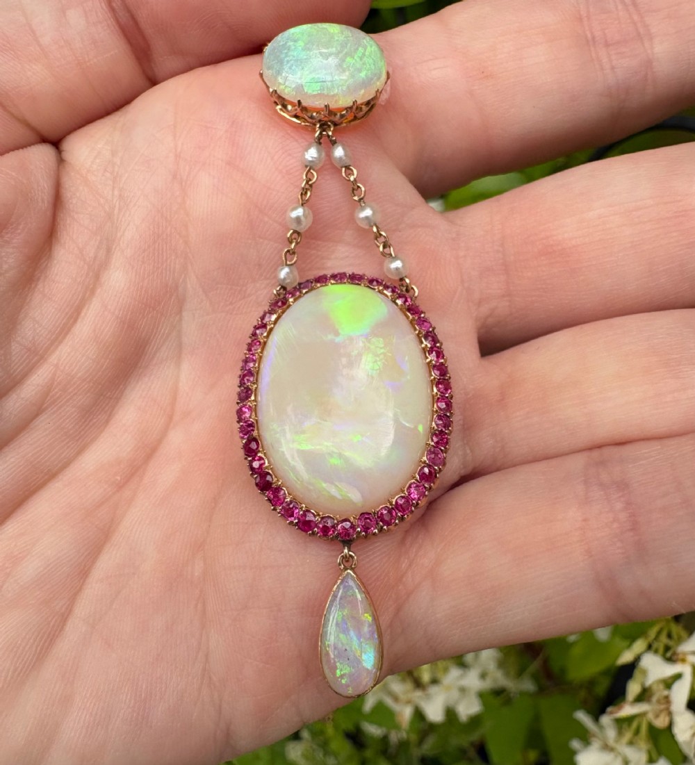 An antique Victorian 15ct yellow gold lavalier pendant set with two oval cabochon opals, a halo of rubies, and seed pearl connectors. View VI