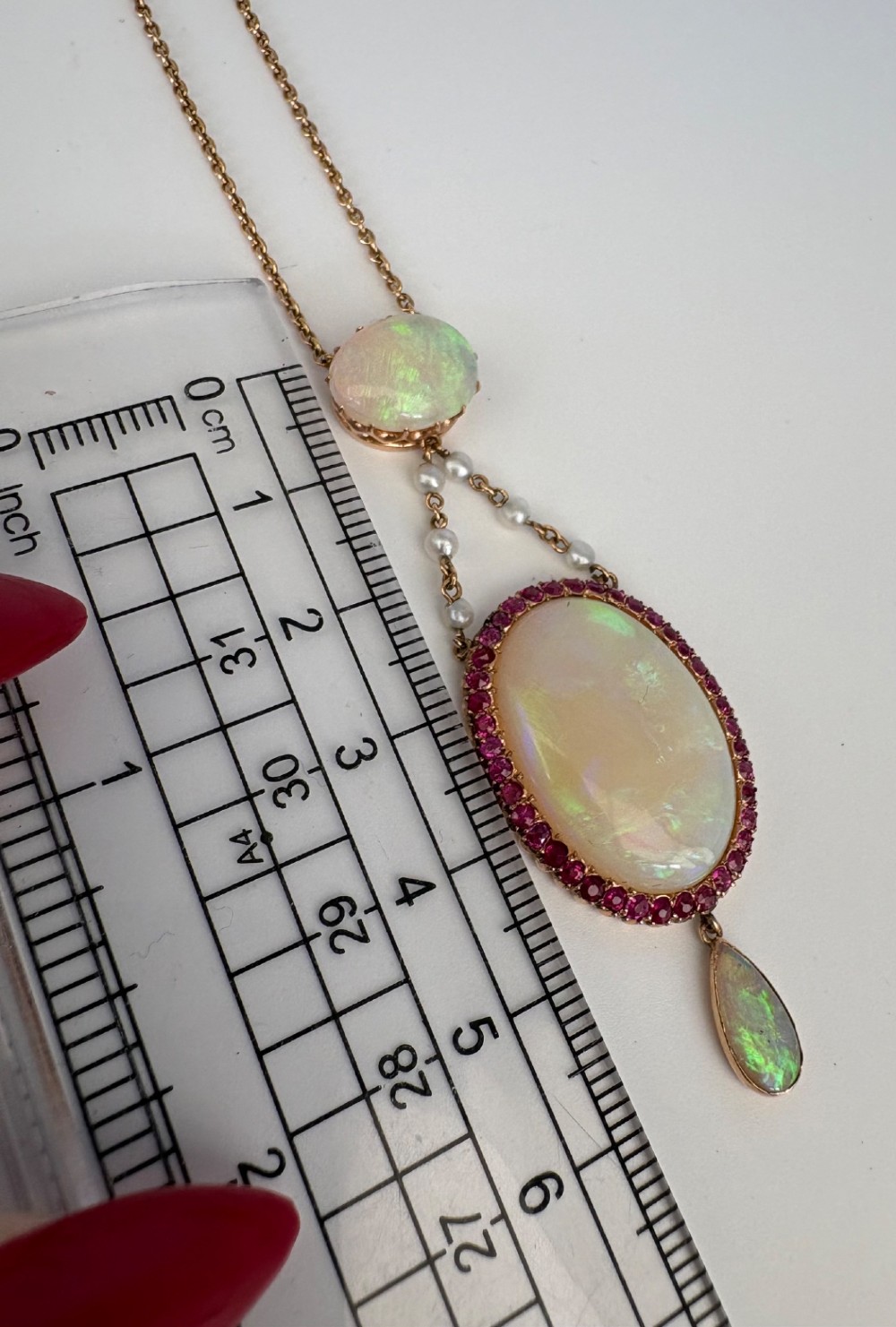 An antique Victorian 15ct yellow gold lavalier pendant set with two oval cabochon opals, a halo of rubies, and seed pearl connectors. View X
