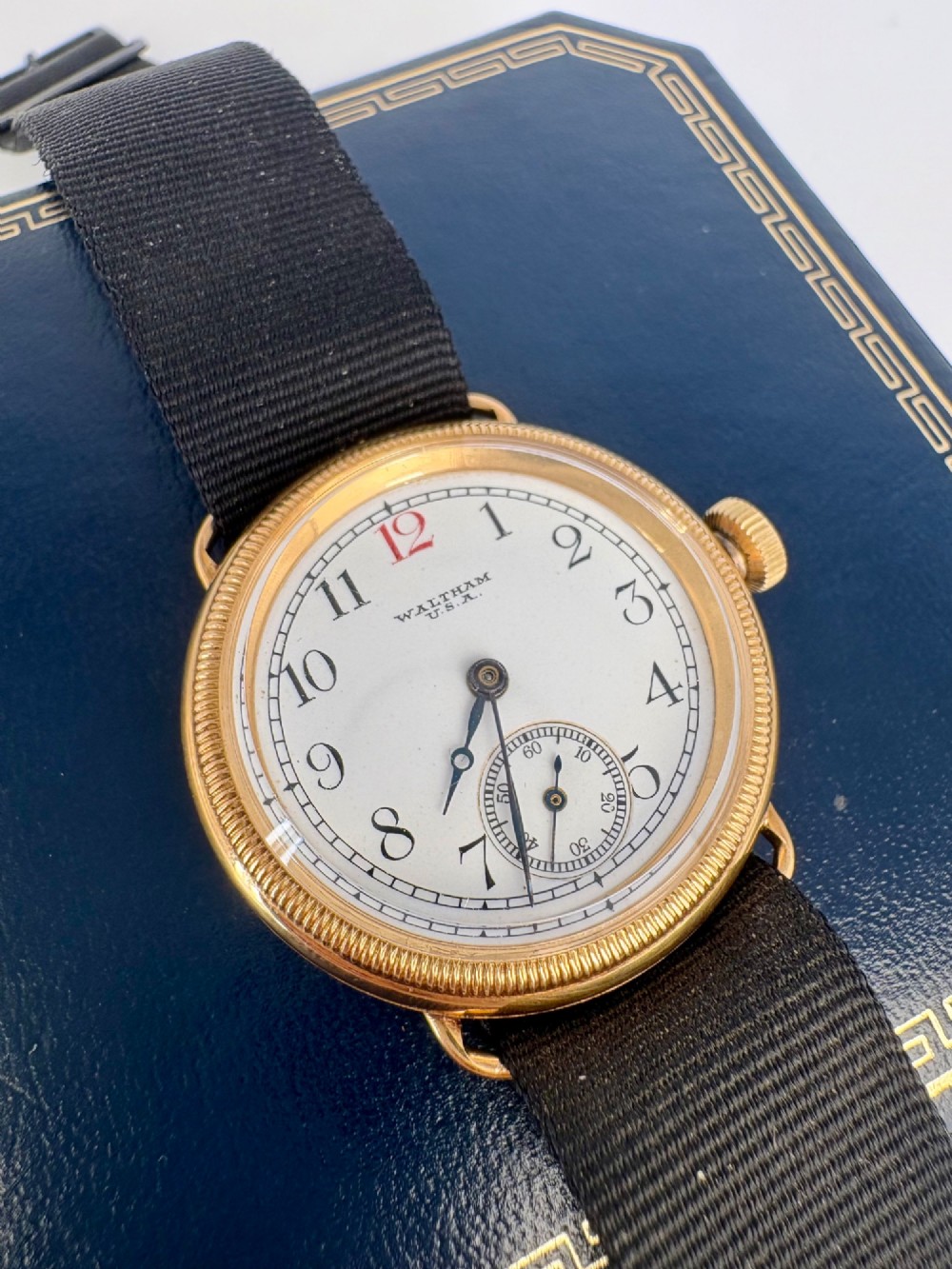 A 1920s 18ct gold Waltham wristwatch with a white enamel dial, black Arabic numerals, a red 12 marker, and a black woven strap.