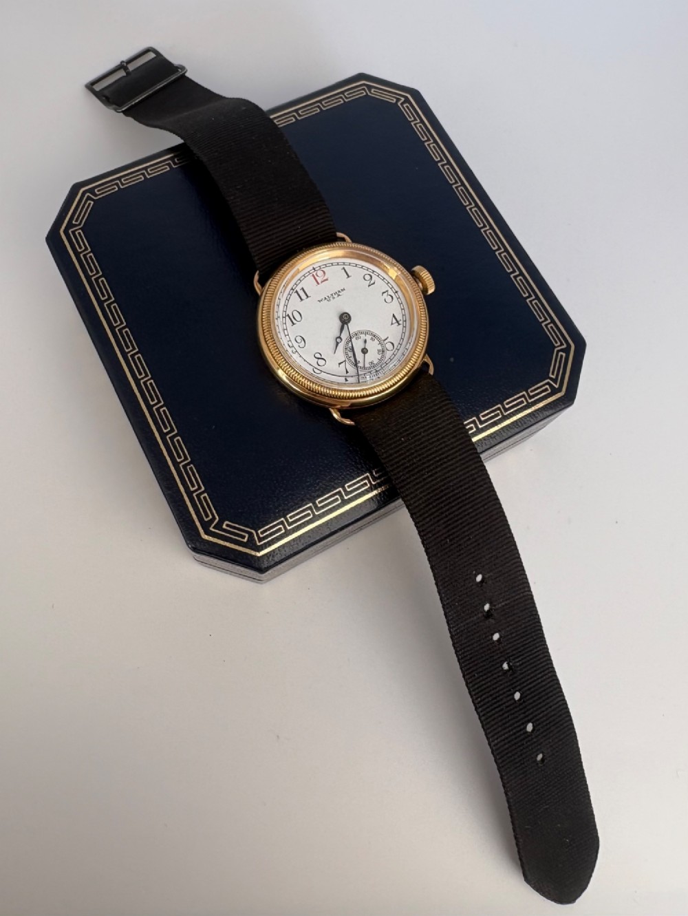A 1920s 18ct gold Waltham wristwatch with a white enamel dial, black Arabic numerals, a red 12 marker, and a black woven strap. View VI