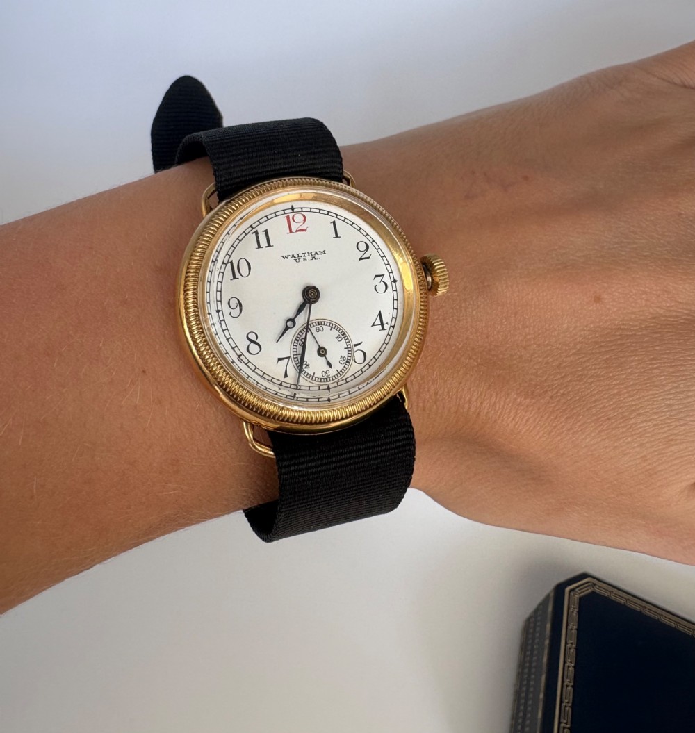 A 1920s 18ct gold Waltham wristwatch with a white enamel dial, black Arabic numerals, a red 12 marker, and a black woven strap. View X