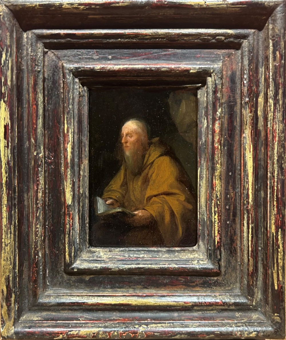 An antique oil painting depicting an elderly bearded hermit in ochre robes reading an open book, presented within a distressed gilded and dark red moulded wooden frame.