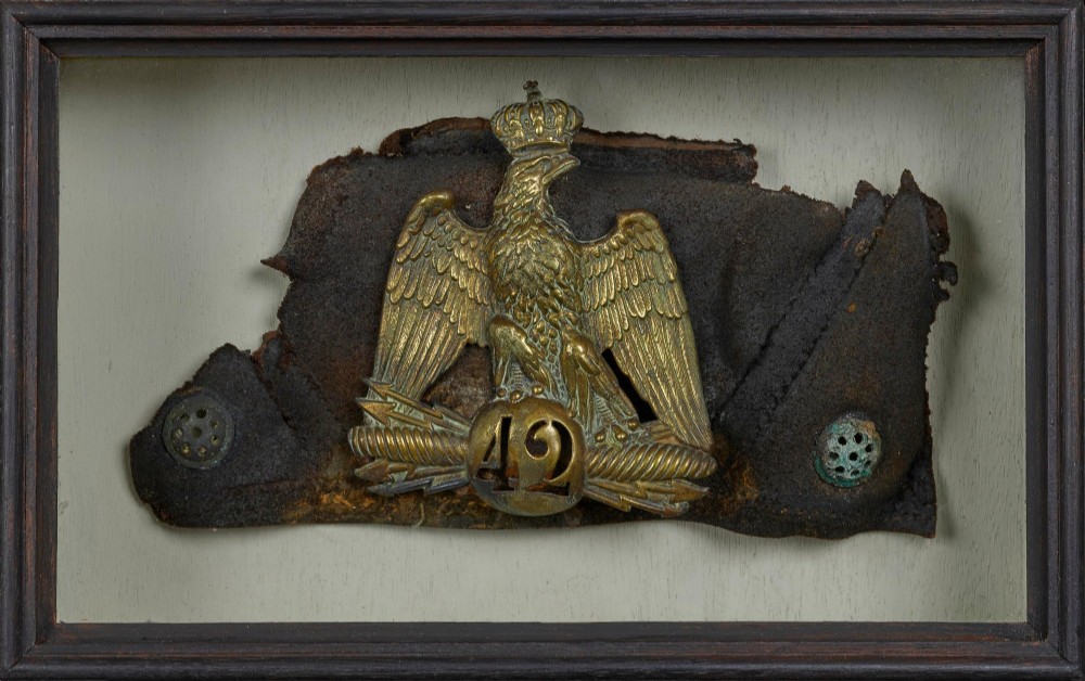 A framed shadow box displaying a stamped brass Napoleonic imperial eagle shako plate bearing the number 42 mounted on a distressed dark leather hat fragment.