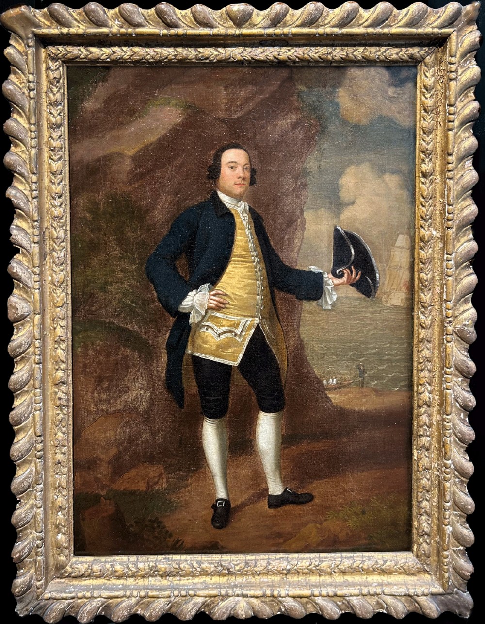 An 18th-century oil portrait of a gentleman in a blue coat and gold waistcoat holding a tricorn hat, framed in a carved giltwood frame.