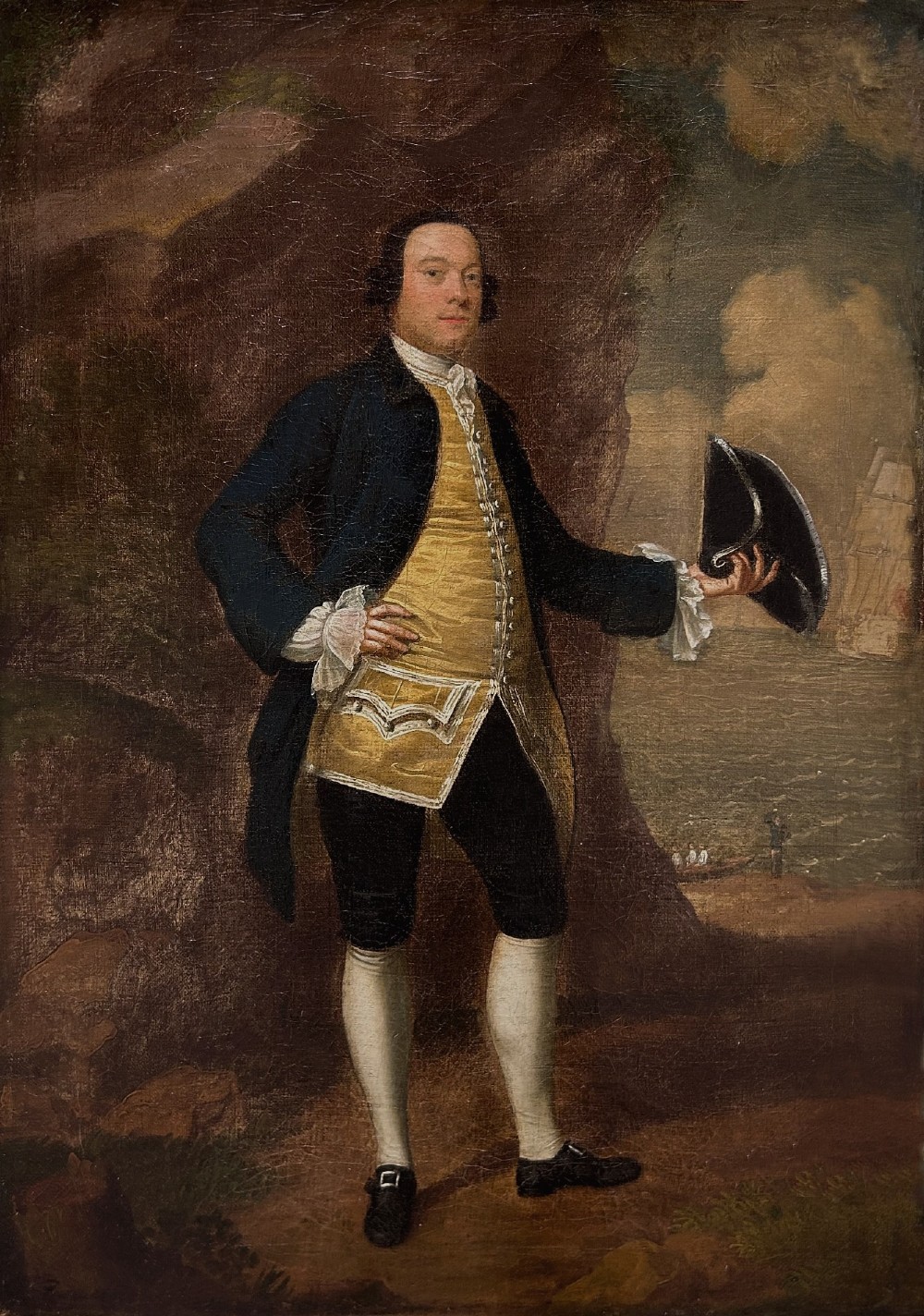 An 18th-century oil portrait of a gentleman in a blue coat and gold waistcoat holding a tricorn hat, framed in a carved giltwood frame. View II