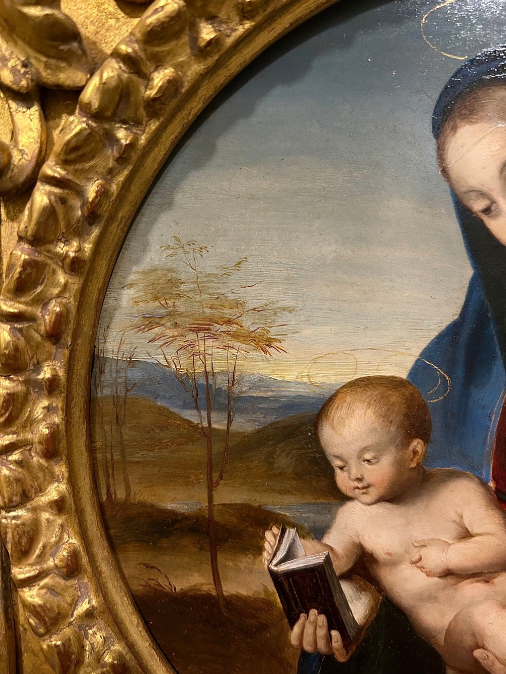 A circular oil painting depicting the Virgin Mary in a blue cloak holding the Christ Child, enclosed within an ornate rectangular carved giltwood frame. View V