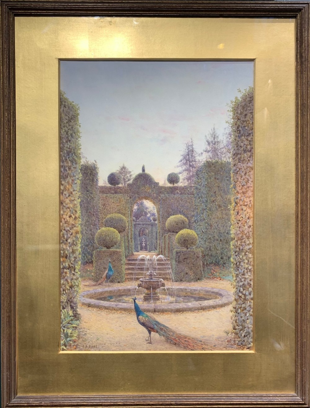A framed watercolour painting depicting a formal garden with manicured topiary, a central stone fountain, and peacocks, set within a wide gold mount and dark wood frame.