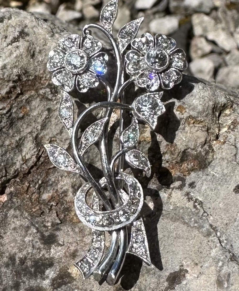A platinum floral spray brooch featuring two diamond-set daisy blooms, leaves, and a ribbon bow tie.