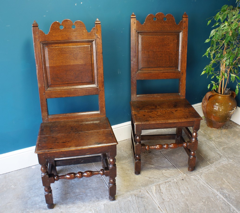 A pair of 17th-century dark oak Lancashire backstool chairs with panelled backs, shaped cresting rails, and turned front legs.