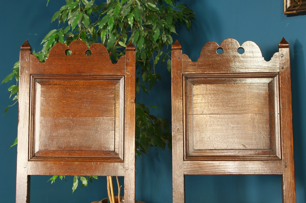 A pair of 17th-century dark oak Lancashire backstool chairs with panelled backs, shaped cresting rails, and turned front legs. View V