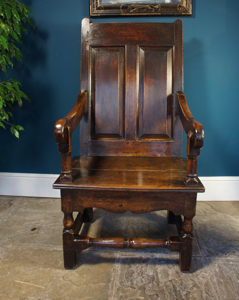 An 18th century dark oak armchair featuring a double-panelled backrest, shaped open arms, and turned front legs connected by a stretcher.