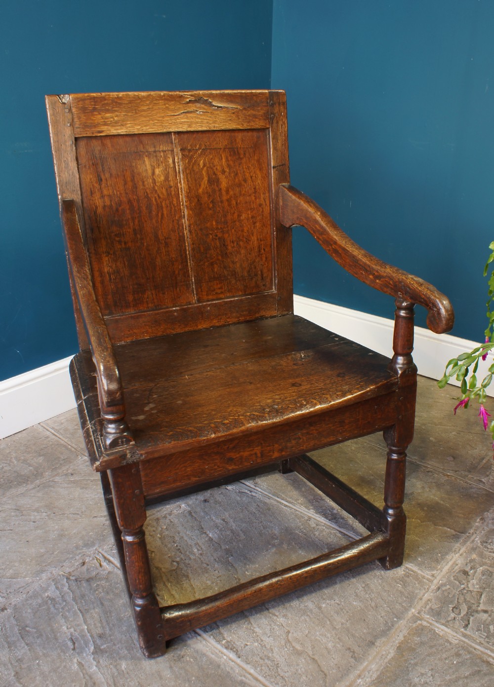 A 17th-century dark oak wainscot armchair featuring a solid panelled back, curved arms, turned front legs, and square stretchers.