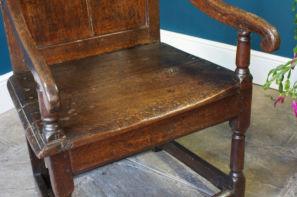 A 17th-century dark oak wainscot armchair featuring a solid panelled back, curved arms, turned front legs, and square stretchers. View II