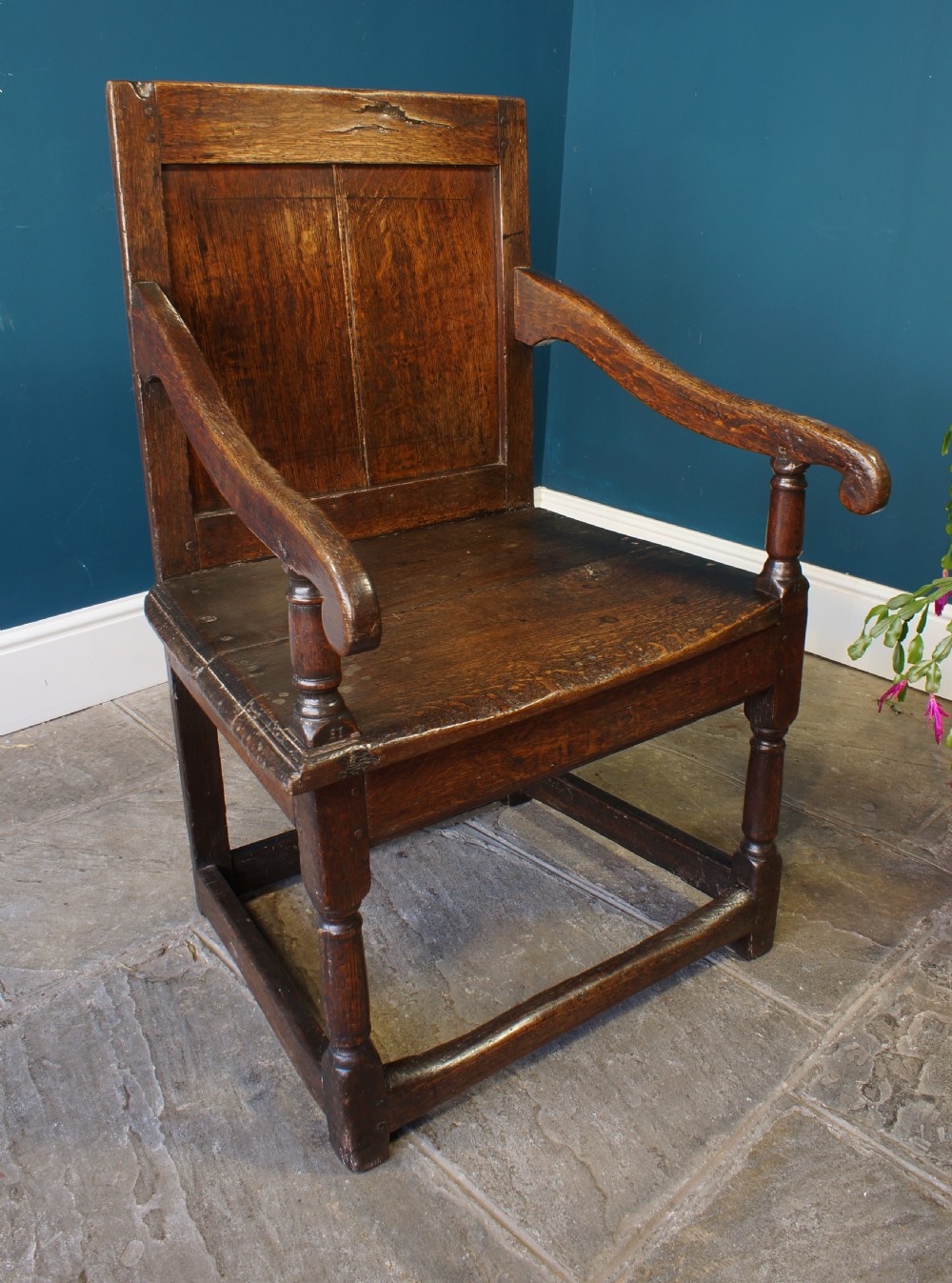 A 17th-century dark oak wainscot armchair featuring a solid panelled back, curved arms, turned front legs, and square stretchers. View XI