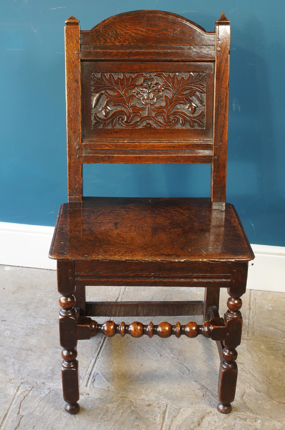 A dark oak 17th-century side chair with a carved floral back panel, solid plank seat, and bobbin-turned front stretcher.