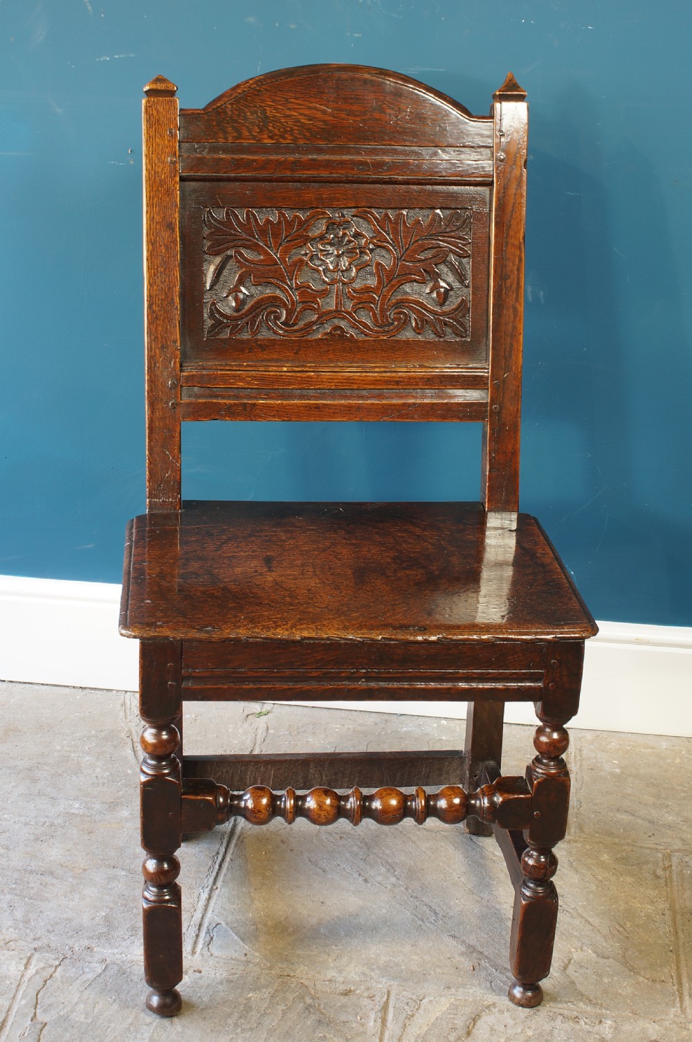 A dark oak 17th-century side chair with a carved floral back panel, solid plank seat, and bobbin-turned front stretcher. View V