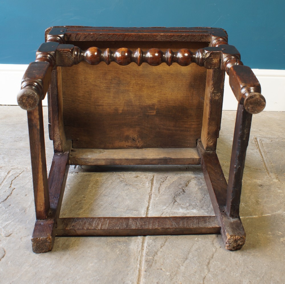 A dark oak 17th-century side chair with a carved floral back panel, solid plank seat, and bobbin-turned front stretcher. View VII