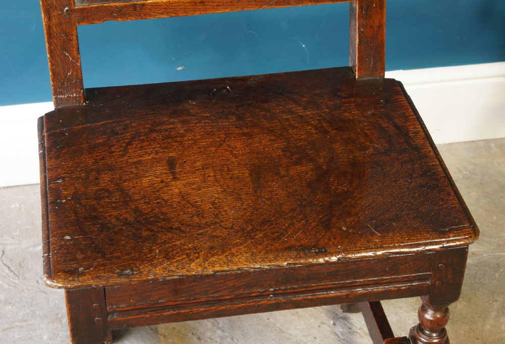A dark oak 17th-century side chair with a carved floral back panel, solid plank seat, and bobbin-turned front stretcher. View X