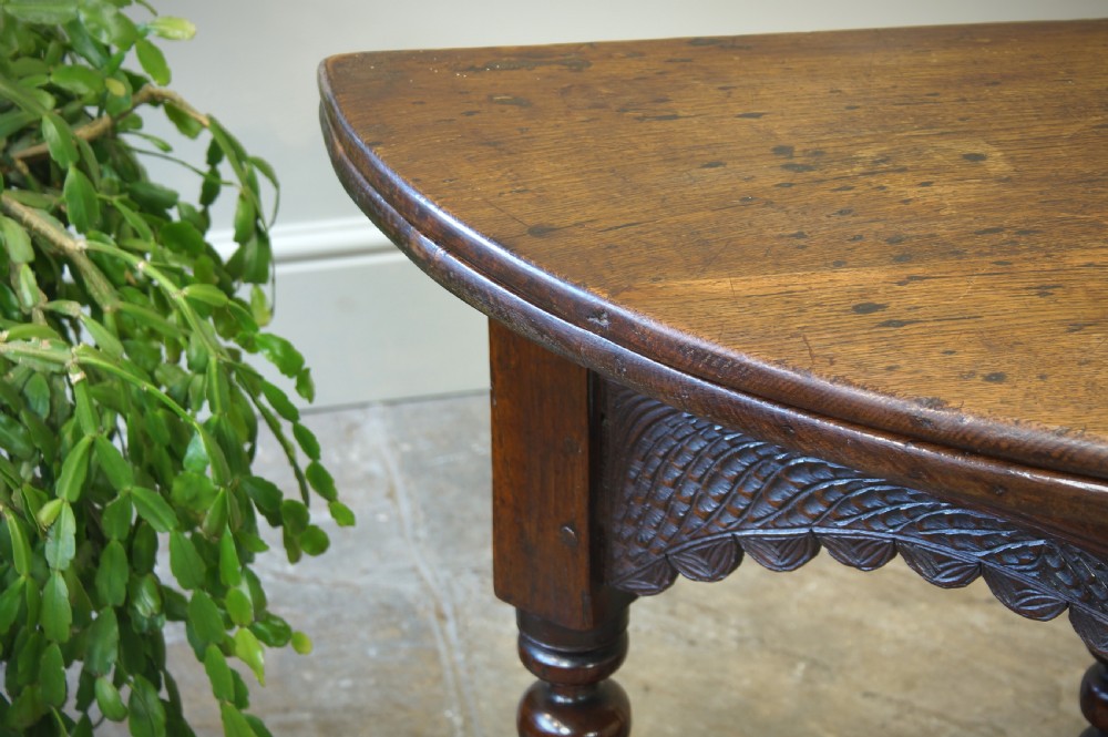 A 17th-century oak credence table featuring a semi-circular fold-over top, hand-carved frieze, turned legs, and low perimeter stretchers. View V