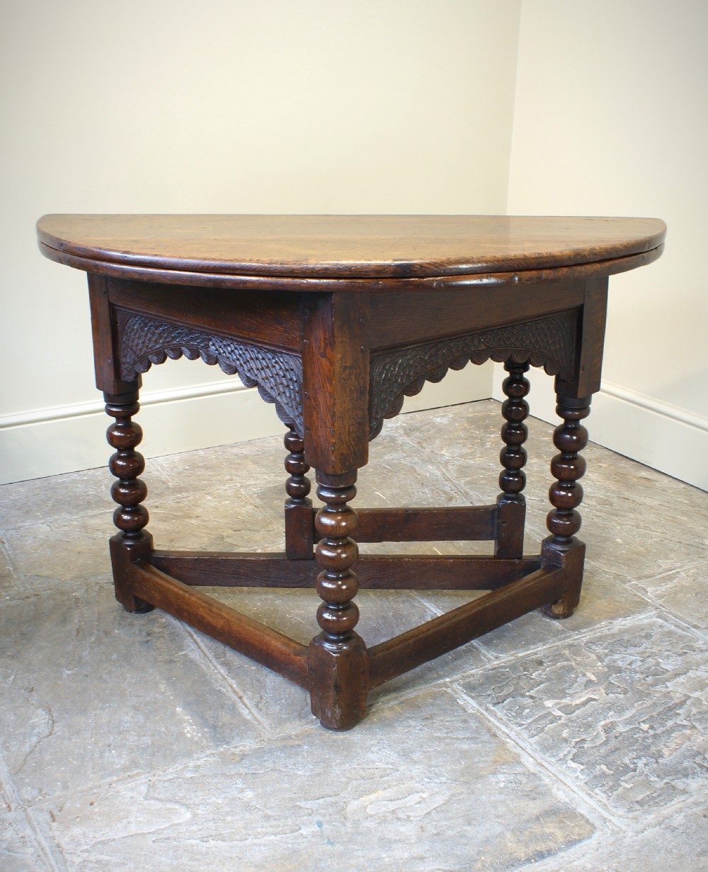 A 17th-century oak credence table featuring a semi-circular fold-over top, hand-carved frieze, turned legs, and low perimeter stretchers. View VI