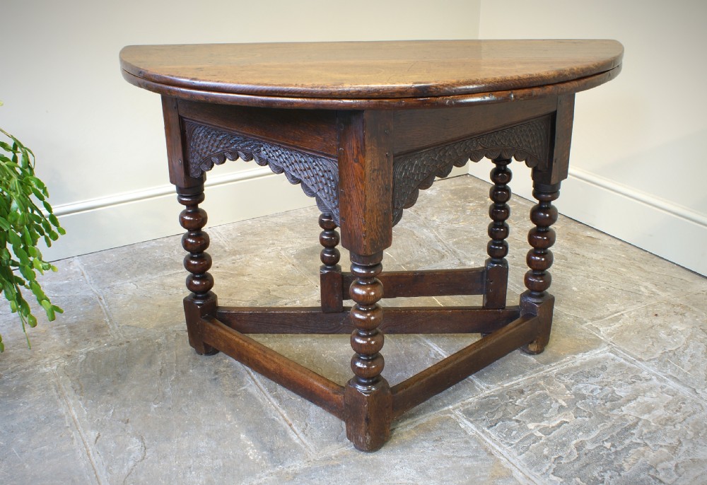 A 17th-century oak credence table featuring a semi-circular fold-over top, hand-carved frieze, turned legs, and low perimeter stretchers. View VII