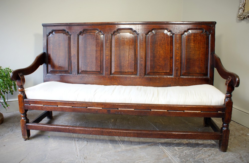 An early eighteenth-century dark oak settle featuring a five-panel shaped backrest, curved armrests, turned front legs, and a cream upholstered seat cushion.