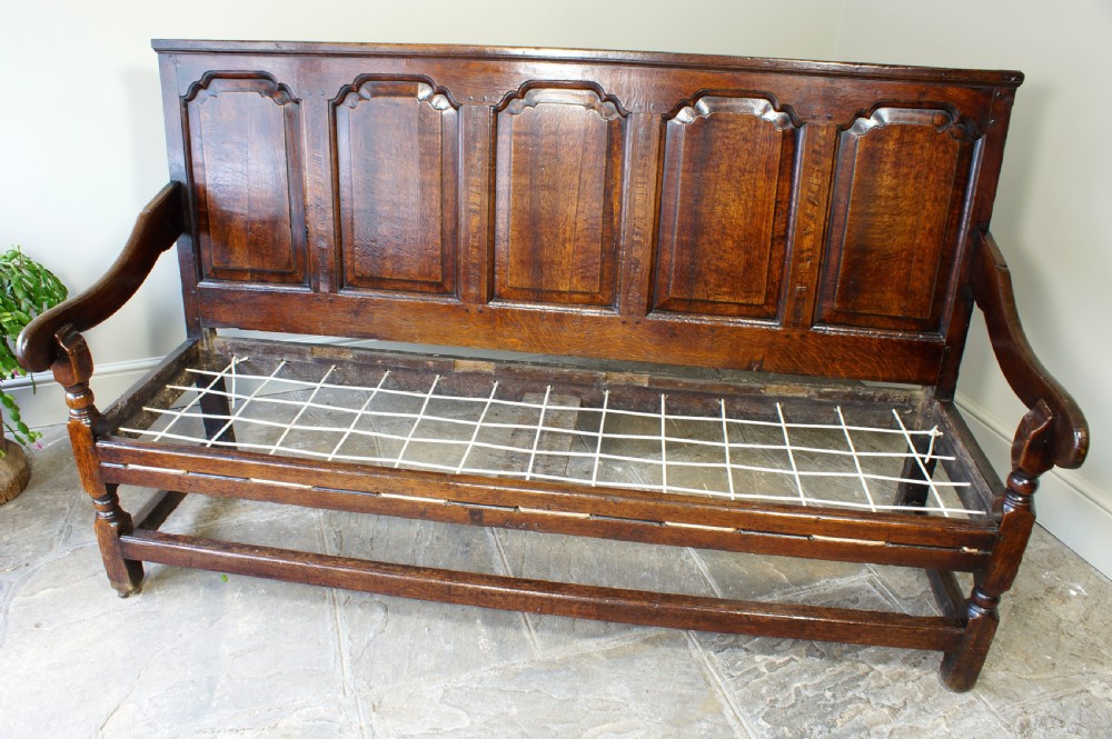 An early eighteenth-century dark oak settle featuring a five-panel shaped backrest, curved armrests, turned front legs, and a cream upholstered seat cushion. View II