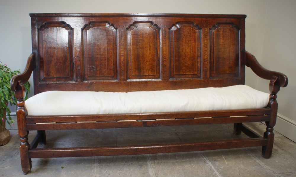 An early eighteenth-century dark oak settle featuring a five-panel shaped backrest, curved armrests, turned front legs, and a cream upholstered seat cushion. View III