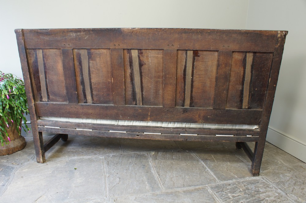 An early eighteenth-century dark oak settle featuring a five-panel shaped backrest, curved armrests, turned front legs, and a cream upholstered seat cushion. View V