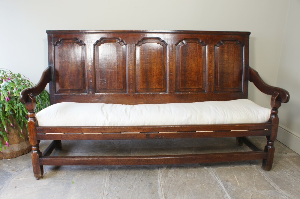 An early eighteenth-century dark oak settle featuring a five-panel shaped backrest, curved armrests, turned front legs, and a cream upholstered seat cushion. View VI