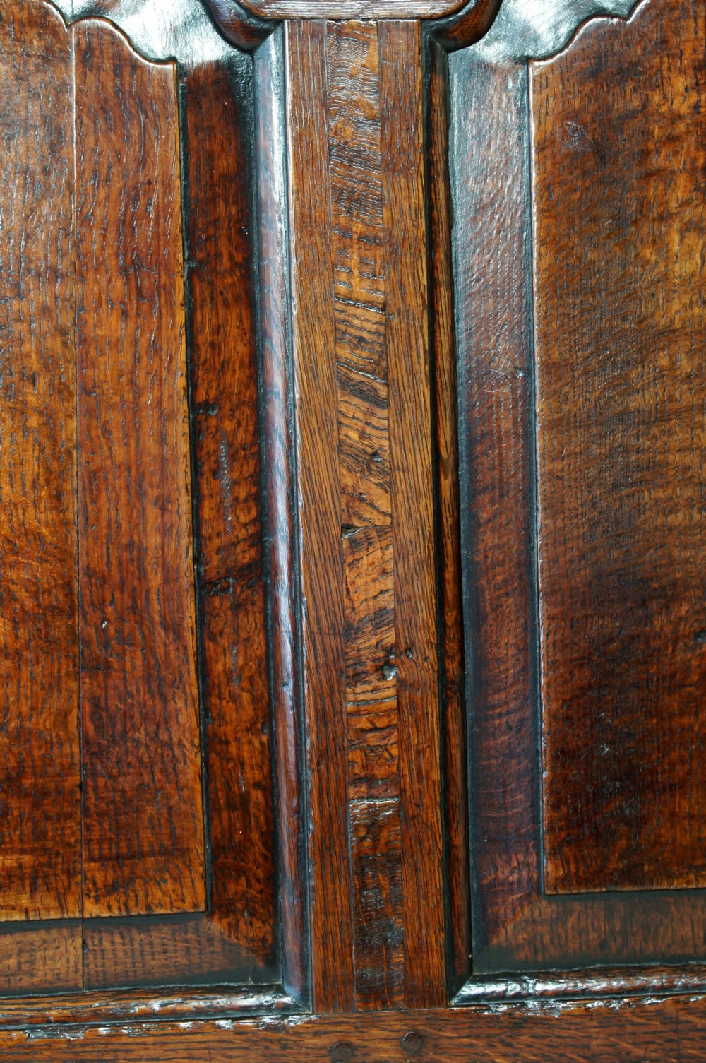 An early eighteenth-century dark oak settle featuring a five-panel shaped backrest, curved armrests, turned front legs, and a cream upholstered seat cushion. View VII
