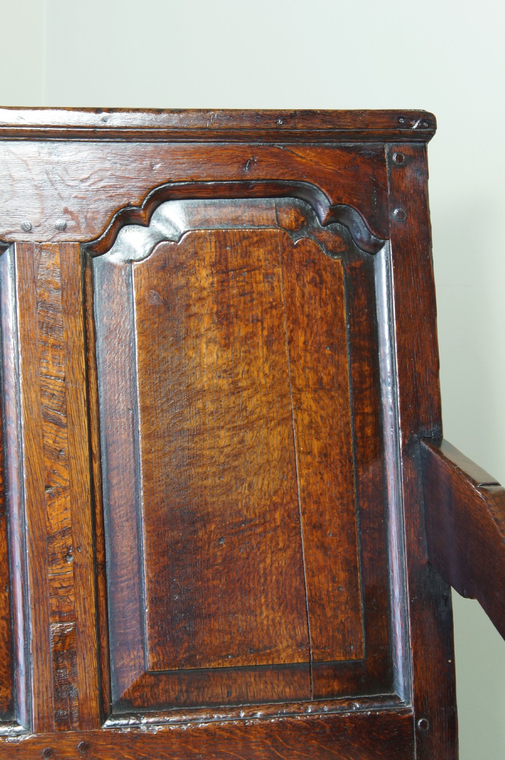 An early eighteenth-century dark oak settle featuring a five-panel shaped backrest, curved armrests, turned front legs, and a cream upholstered seat cushion. View IX