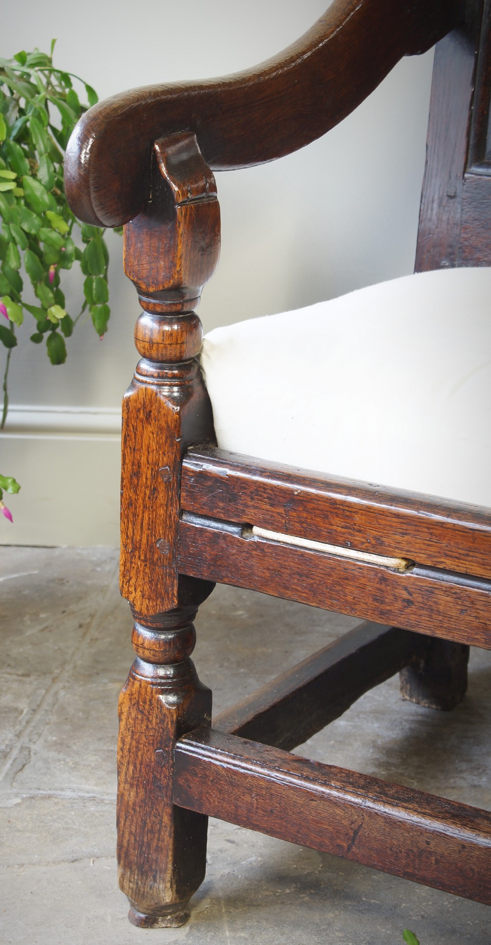 An early eighteenth-century dark oak settle featuring a five-panel shaped backrest, curved armrests, turned front legs, and a cream upholstered seat cushion. View X