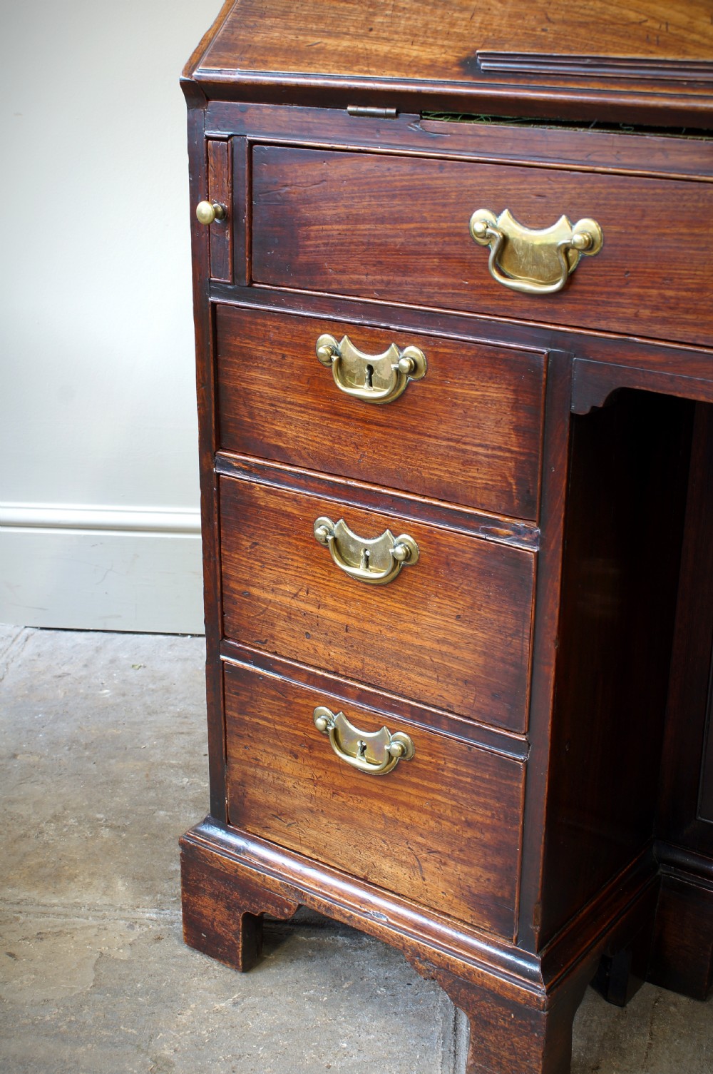 An early Georgian mahogany kneehole bureau featuring a slanted fall-front top lid, brass drop handles, multiple drawers, and a central recessed cupboard on bracket feet. View III