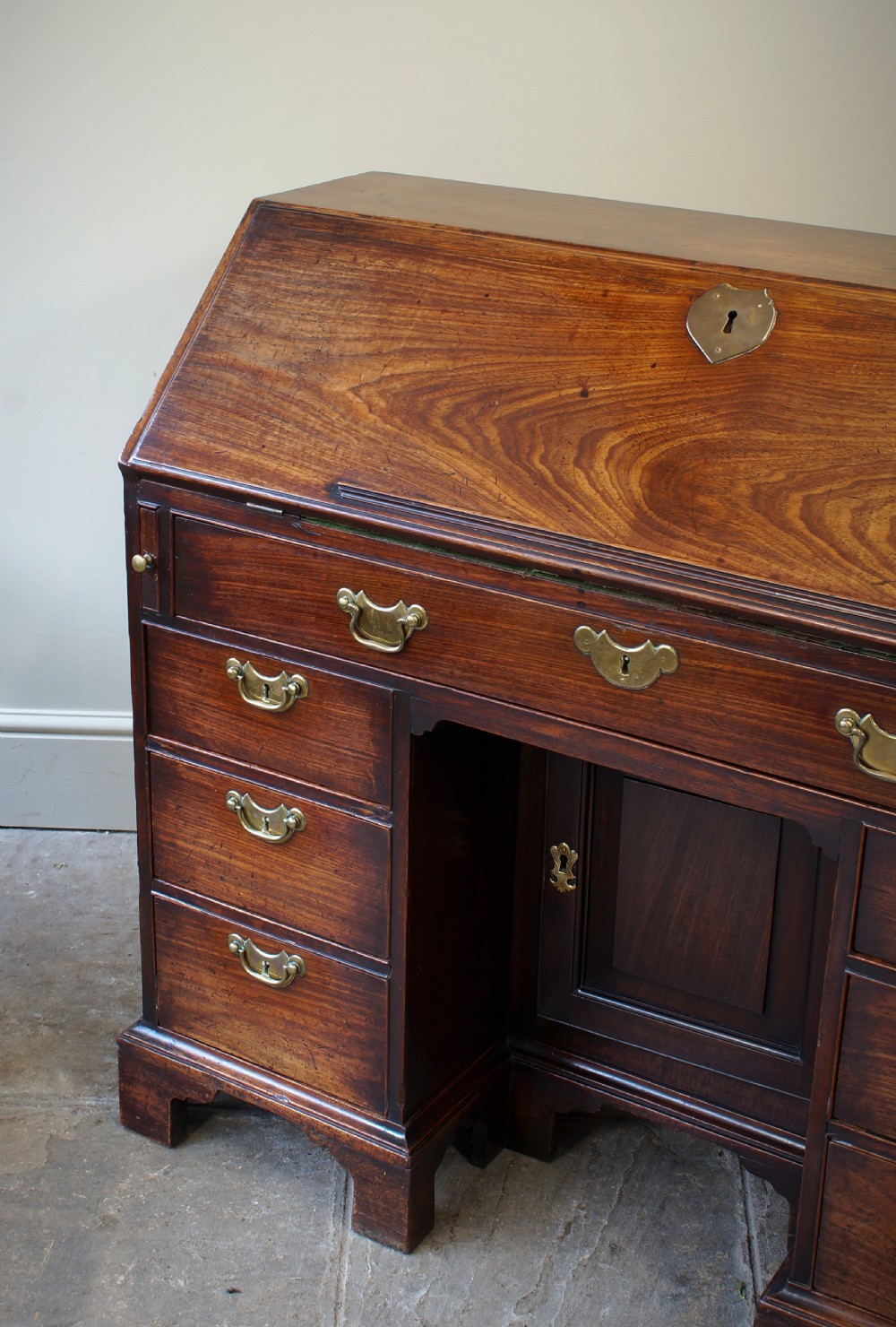 An early Georgian mahogany kneehole bureau featuring a slanted fall-front top lid, brass drop handles, multiple drawers, and a central recessed cupboard on bracket feet. View V