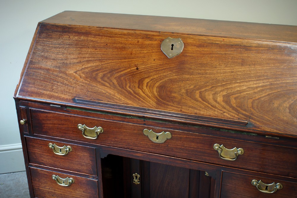 An early Georgian mahogany kneehole bureau featuring a slanted fall-front top lid, brass drop handles, multiple drawers, and a central recessed cupboard on bracket feet. View VI