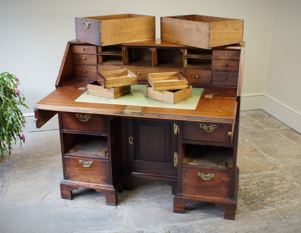An early Georgian mahogany kneehole bureau featuring a slanted fall-front top lid, brass drop handles, multiple drawers, and a central recessed cupboard on bracket feet. View VIII