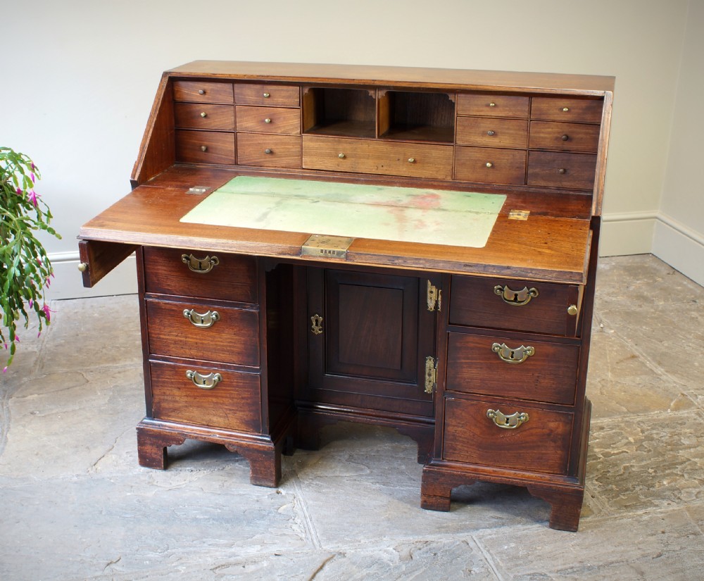An early Georgian mahogany kneehole bureau featuring a slanted fall-front top lid, brass drop handles, multiple drawers, and a central recessed cupboard on bracket feet. View IX