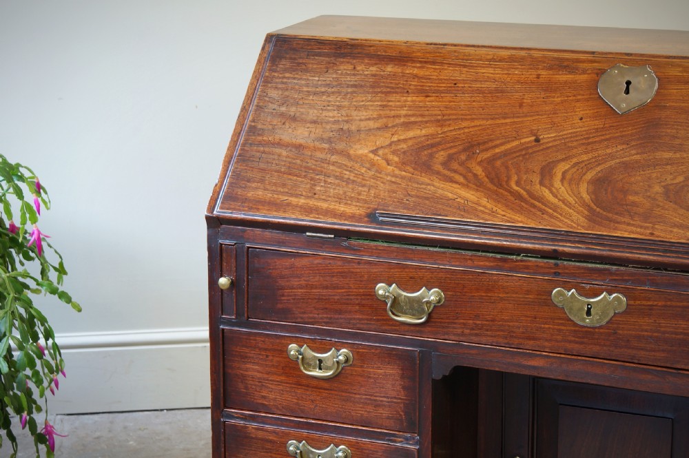 An early Georgian mahogany kneehole bureau featuring a slanted fall-front top lid, brass drop handles, multiple drawers, and a central recessed cupboard on bracket feet. View X