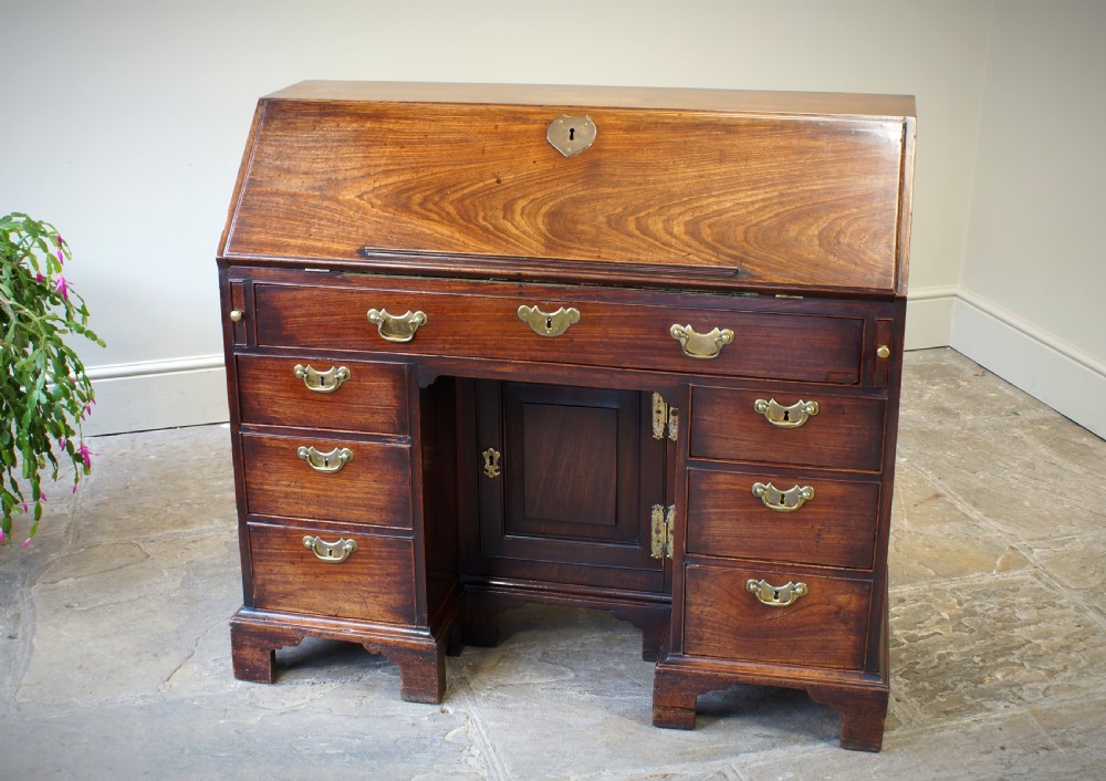 An early Georgian mahogany kneehole bureau featuring a slanted fall-front top lid, brass drop handles, multiple drawers, and a central recessed cupboard on bracket feet.