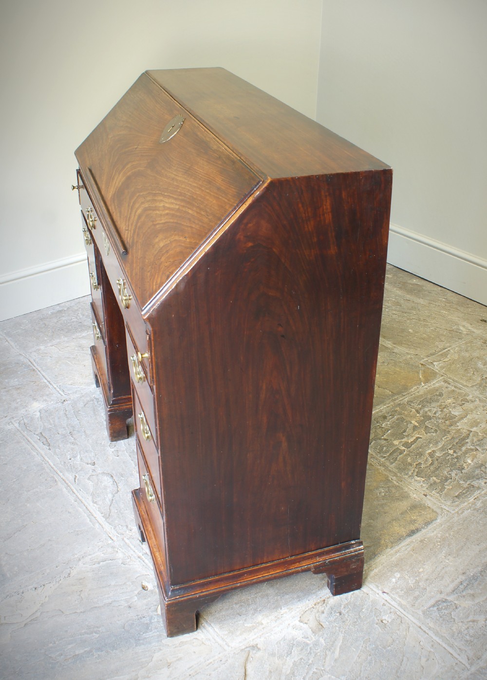 An early Georgian mahogany kneehole bureau featuring a slanted fall-front top lid, brass drop handles, multiple drawers, and a central recessed cupboard on bracket feet. View XII
