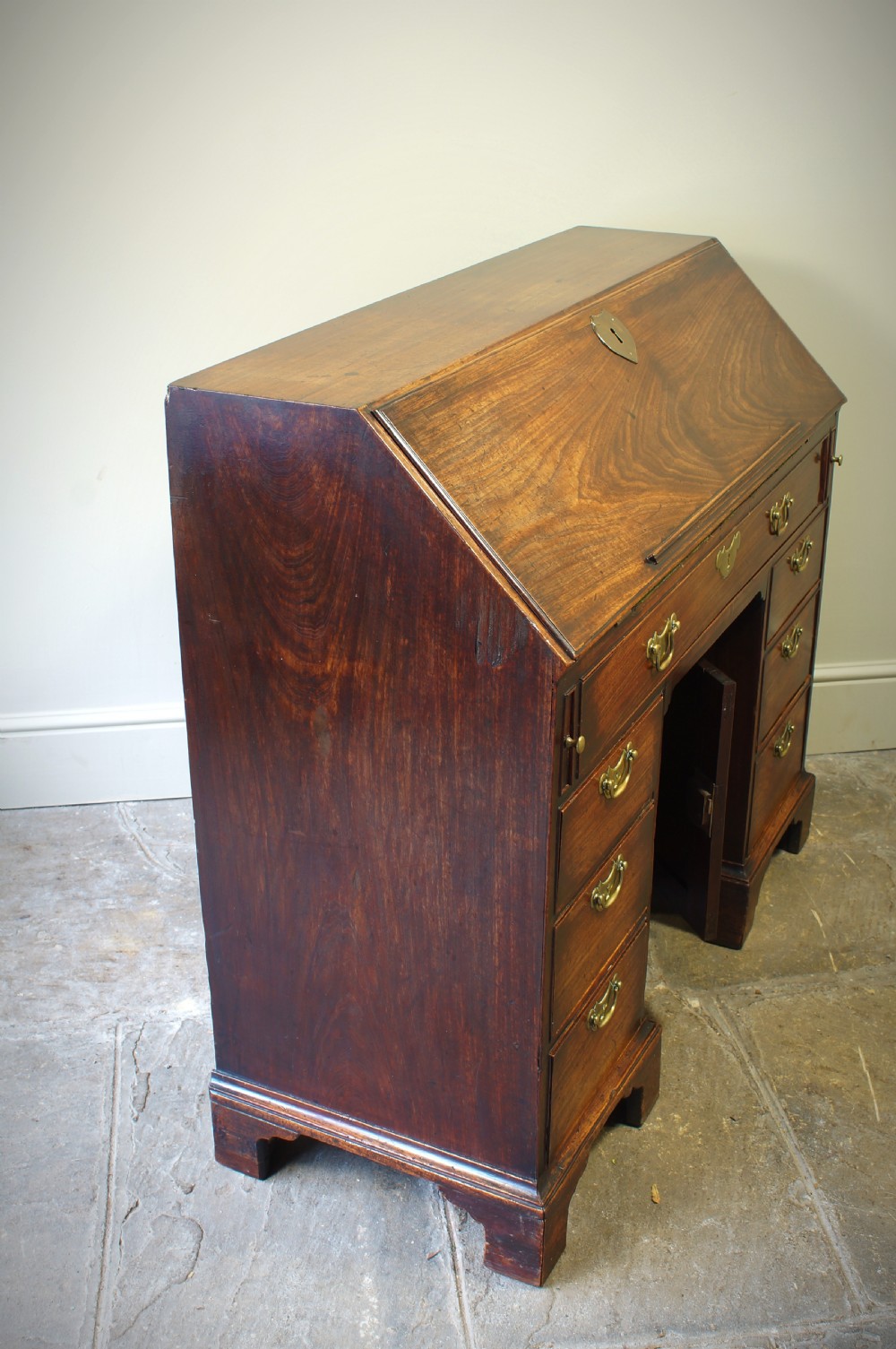 An early Georgian mahogany kneehole bureau featuring a slanted fall-front top lid, brass drop handles, multiple drawers, and a central recessed cupboard on bracket feet. View XIV