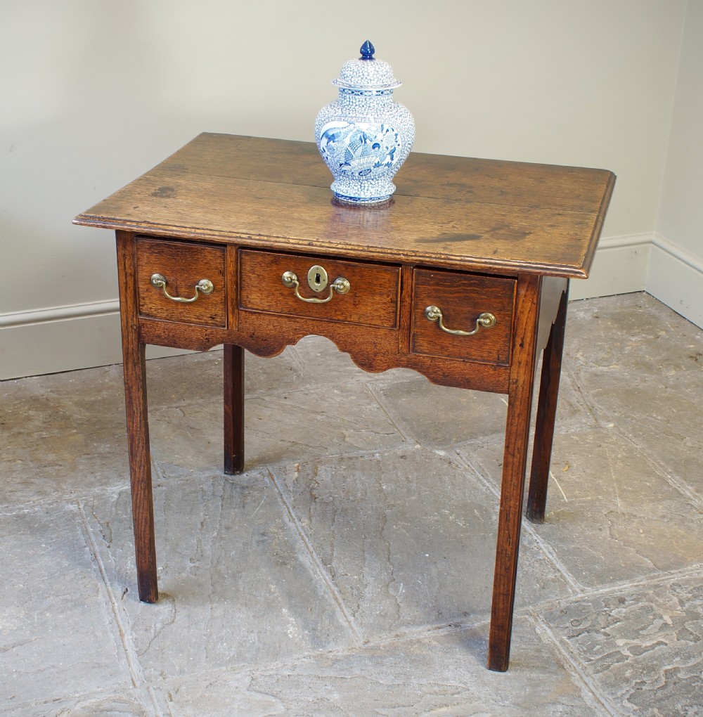 A George III oak lowboy side table featuring three drawers with brass swan-neck handles, a shaped apron, and four square legs.