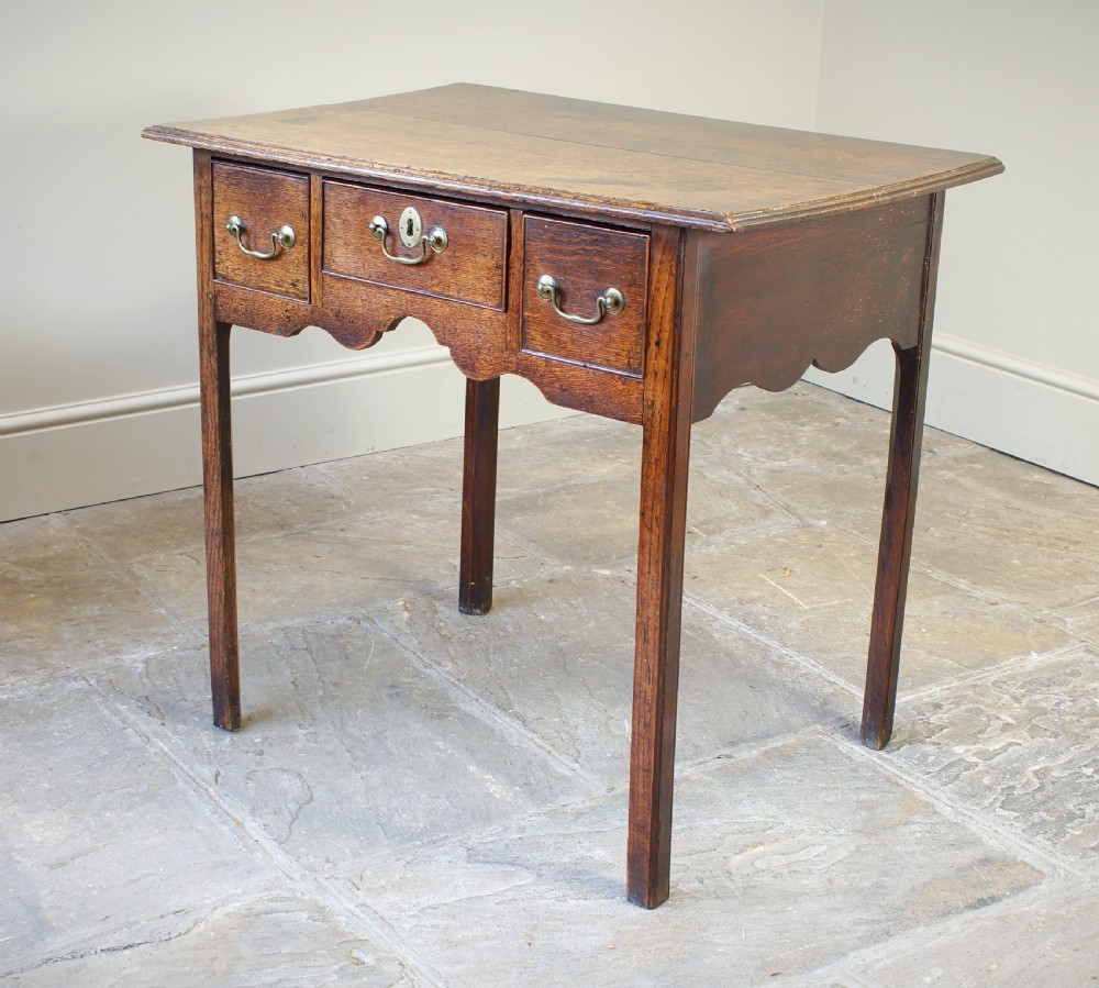 A George III oak lowboy side table featuring three drawers with brass swan-neck handles, a shaped apron, and four square legs. View II