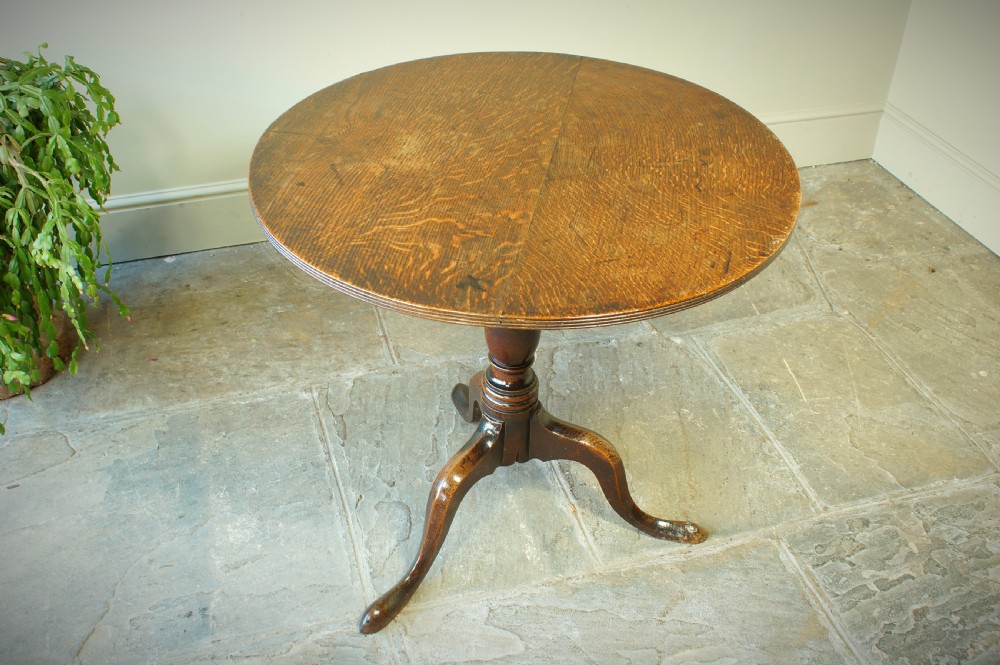 A Georgian oak tilt-top tripod table with a circular top shown in a vertical position, supported by a turned column and three splayed cabriole legs. View II
