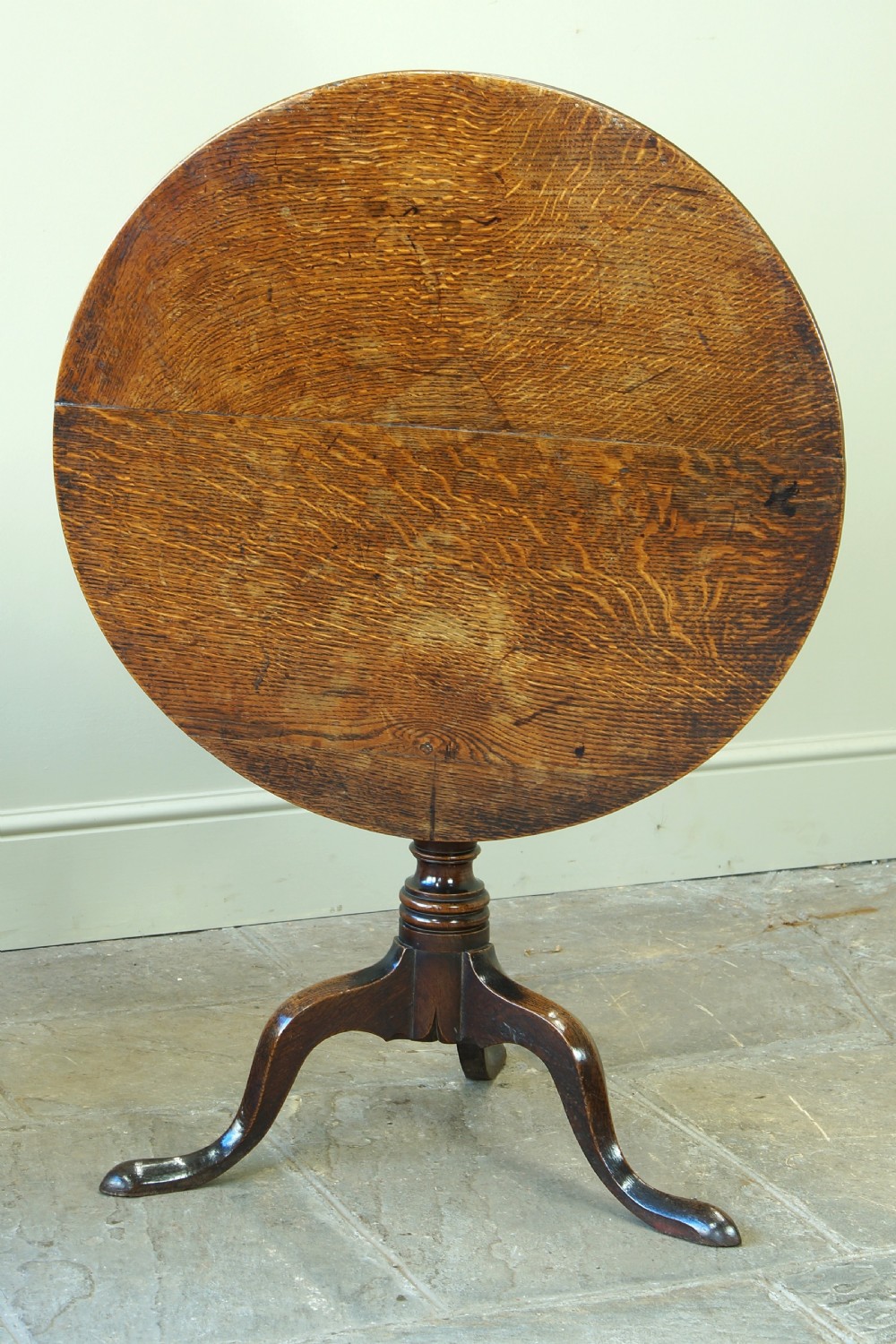 A Georgian oak tilt-top tripod table with a circular top shown in a vertical position, supported by a turned column and three splayed cabriole legs. View IX