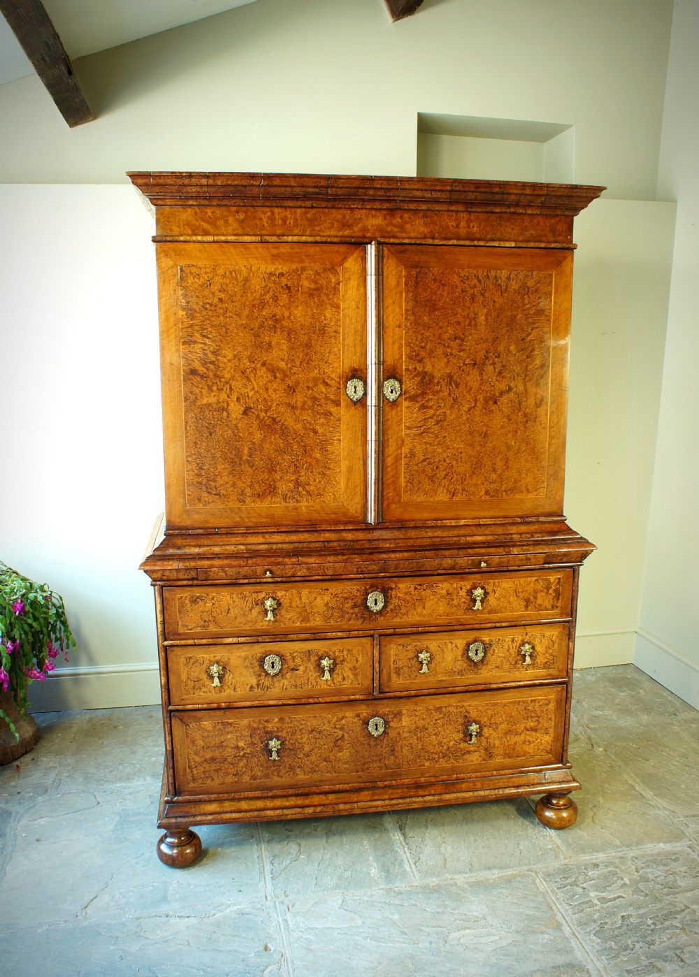 A William and Mary style cabinet on chest featuring double doors with burr amboyna wood veneers, brass keyhole escutcheons, multiple lower drawers, and compressed bun feet.