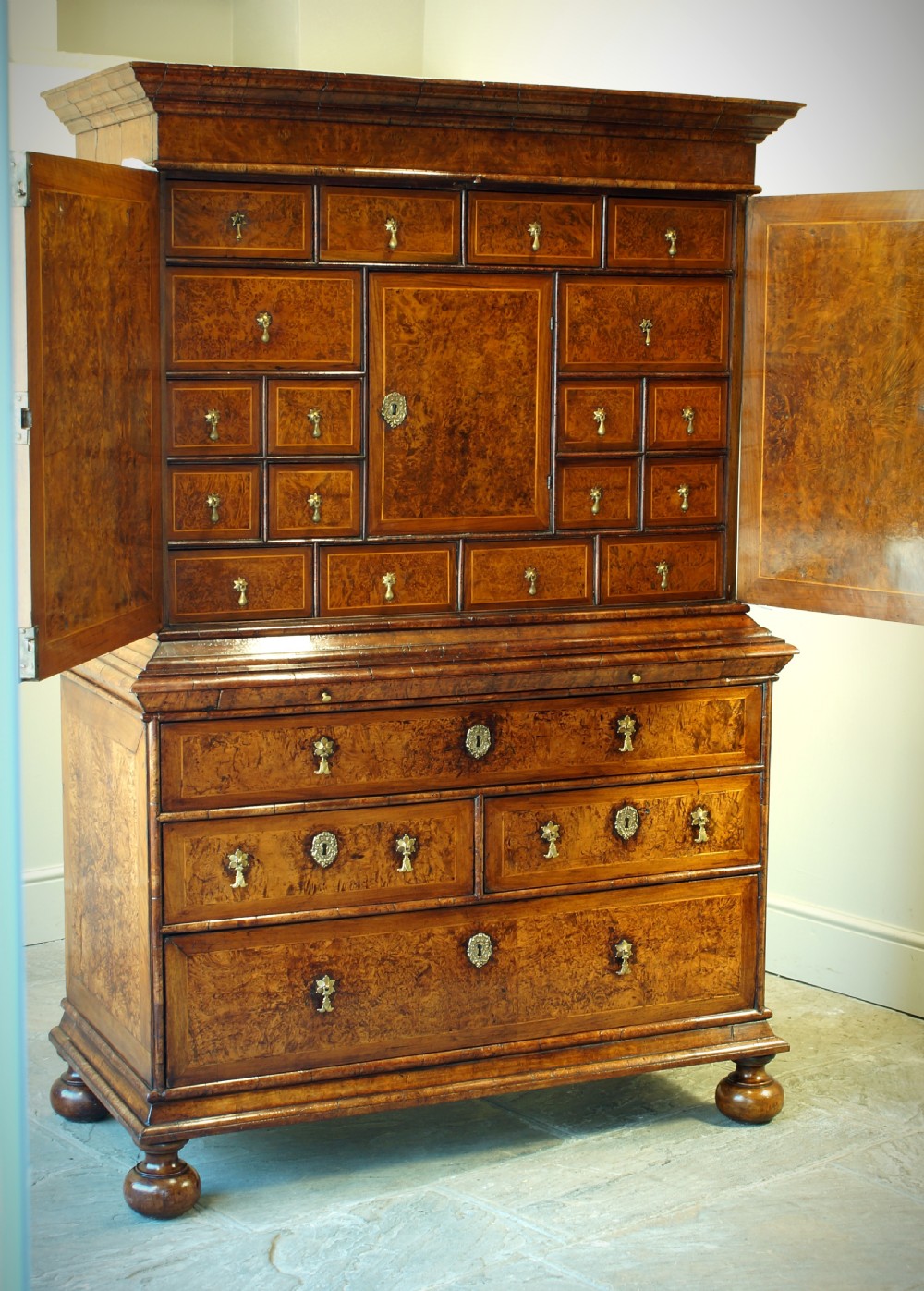 A William and Mary style cabinet on chest featuring double doors with burr amboyna wood veneers, brass keyhole escutcheons, multiple lower drawers, and compressed bun feet. View II