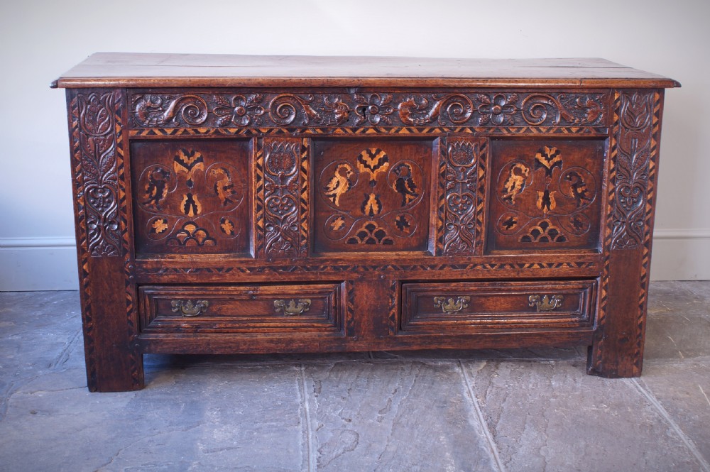A 17th-century dark oak mule chest featuring carved friezes, three inlaid marquetry front panels with bird motifs, and two lower drawers with brass drop handles.